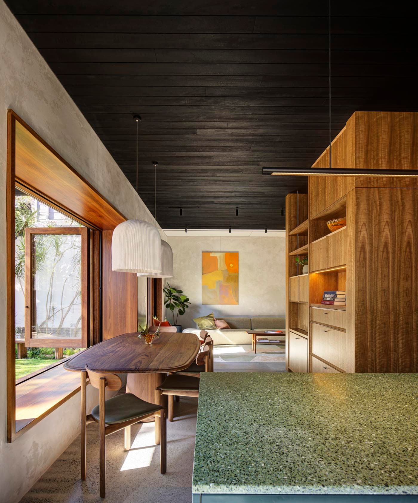 Modern dining and living space by an Inner West architect, featuring a wooden table, chairs, large window seat, green countertop, wood paneling, pendant lights, and a colorful abstract painting on the wall in the background.